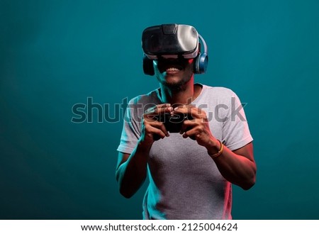 African american guy playing video game with controller and vr headset, enjoying online play with futuristic 3d simulation in studio. Gamer with virtual reality glasses holding joystick.