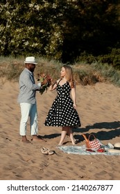 African American Guy Proposes To His Girlfriend On The Beach. He Puts The Ring On Her Hand. A Young Interracial Couple Had A Picnic On The Beach. Romantic Date On A Summer Day 