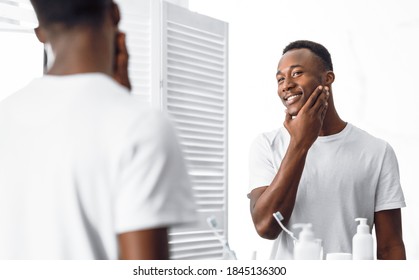 African American Guy Touching Well Shaved Smooth Face After Shaving Routine Standing Near Mirror In Modern Bathroom At Home. Male Beauty And Facial Care. Selective Focus