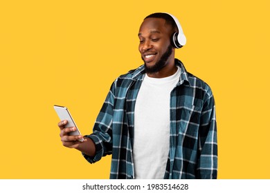 African American Guy Using Phone Wearing Headphones Listening To Music Online Standing In Studio Over Yellow Background. Musical App Advertisement, Favorite Song Concept - Shutterstock ID 1983514628