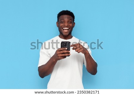 african american guy in white t-shirt uses smartphone and chooses on blue isolated background, man holds phone and presses his finger