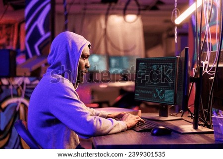 African american hacker coding spyware in abandoned warehouse. Man in hood using computer for breaching data, cracking password and getting access to server database at night