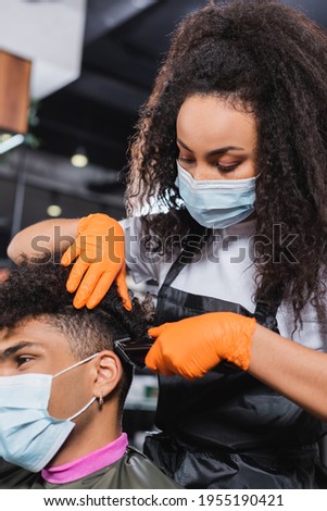 African american hairdresser in medical mask and gloves trimming hair of client on blurred foreground