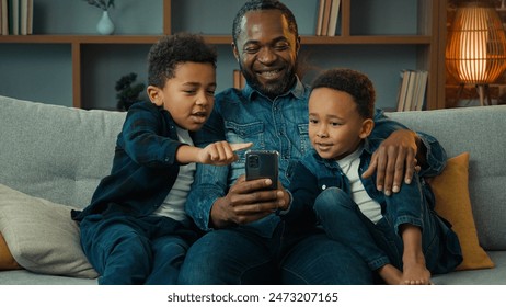 African American happy family father with ethnic sons boys little children siblings kids at home couch sofa using mobile phone together watching funny video looking at smartphone app video call chat - Powered by Shutterstock