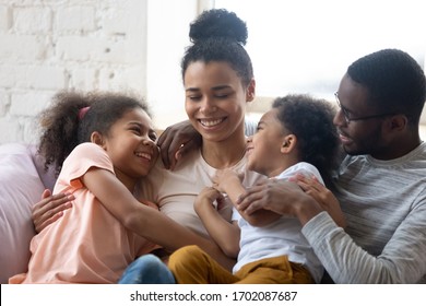 African American Happy Family With Two Kids Enjoy Time Together. Cute Diverse Daughter And Son With Parents Sit On Warm Comfy Couch In Living Room. Young Father And Mother Have Fun With Adorable Kids