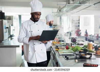 African american head chef with laptop searching for dish recipes on internet while standing in restaurant kitchen. Professional cook wearing cooking uniform using computer to inspire for gourmet meal - Powered by Shutterstock