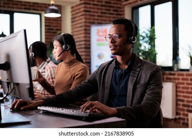 African american helpline employee working at call center reception with multiple monitors. Male operator using telecommunication to help clients at customer service support, remote network.