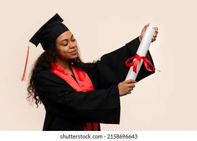 African American High School Student Celebrates Graduation And Rejoices Receiving Certificate Of Education. Dark-skinned Girl In Mortarboard And Graduation Gown Holds Diploma Tied With Red Ribbon