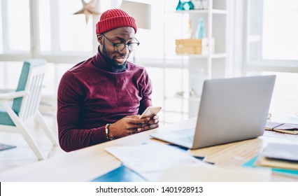 African American Hipster Guy In Spectacles Sending Text Message On Mobile Phone While Working Online Via Netbook At Home,millenial Dark Skinned Man Checking Notification About Transaction On Cellular
