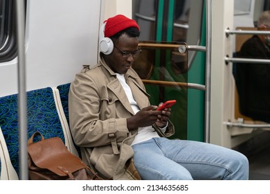 African American Hipster In Trench Coat, Red Hat In Subway Train Thinking Using Cellphone. Bored Black Guy In Glasses Finds Out News, Listening Music With Wireless Headphones In Public Transportation