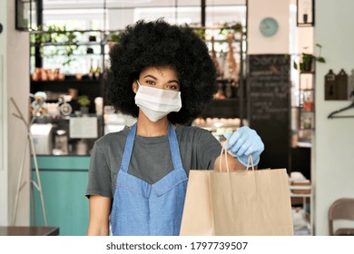 African American hipster waitress wearing face mask and gloves holding takeaway food order in hands giving bag standing in cafe. Coffee shop worker offering takeout safe restaurant take away delivery