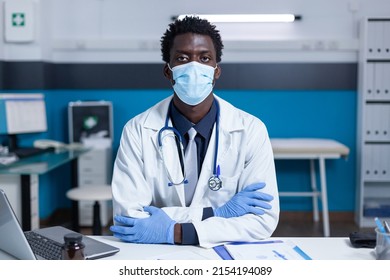 African American Hospital Doctor Sitting At Office Desk While Wearing Facemask Because Of Coronavirus Pandemic. Clinic Healthcare Specialist Medic Sitting At Desk While Wearing Virus Protection Mask