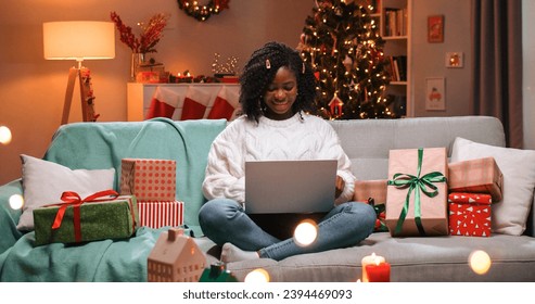 African American joyful beautiful young woman sitting on couch in decorated room with xmas tree and typing on laptop in good mood spending Christmas time on internet. New Year concept. - Powered by Shutterstock