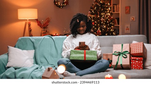 African American joyful beautiful young woman sitting on sofa in decorated house in evening on Christmas Eve with xmas gift box typing on smartphone and smiling. New Year night. Xmas concept - Powered by Shutterstock