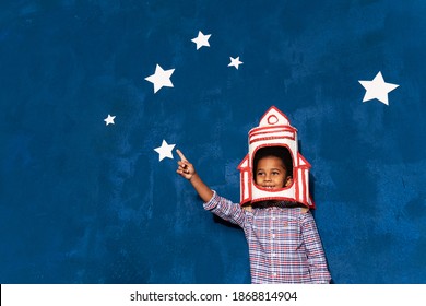African American Kid Boy With Rocket Helmet On Head On Blue Studio Background With White Stars. Portrait Of Smiling Afro Child Playing In Astronaut. Imagination And Childish Dream About Cosmic Space.