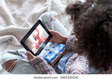 African American Kid Girl Talking To Virtual Friend Indian Child Opening Christmas Gifts During Online Meeting On New Year Holiday By Video Conference Zoom Call Using Tablet Computer Sitting On Bed.