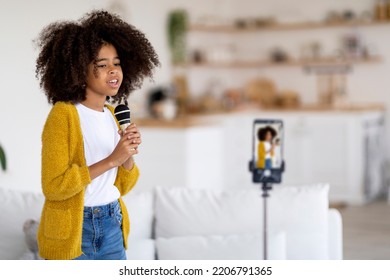 African American Kid Pretty Preteen Girl With Bushy Hair Singing In Front Of Camera, Holding Microphone And Looking At Cell Phone Set On Tripod, Recording Video At Home, Copy Space