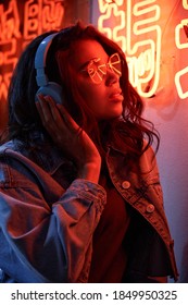 African American Lady Wearing Headphones Listening Music In Neon Light. Cool Funky Young Mixed Race Hipster Listening Trendy Dance Music Stands Near Neon Night Club Sign. Translation: Chicken Laugh