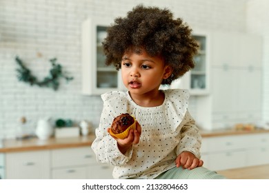 An African American Little Baby With Curly Fluffy Hair In A Dress Eats A Chocolate Muffin With Appetite In The Kitchen At Home.