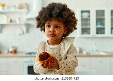 An African American Little Baby With Curly Fluffy Hair In A Dress Eats A Chocolate Muffin With Appetite In The Kitchen At Home.