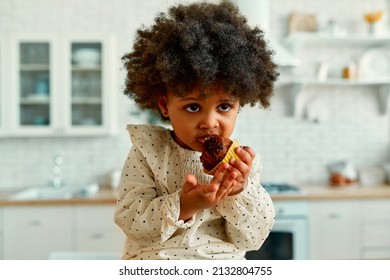 An African American Little Baby With Curly Fluffy Hair In A Dress Eats A Chocolate Muffin With Appetite In The Kitchen At Home.