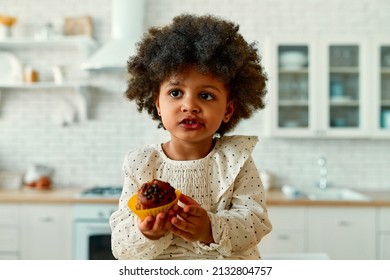 An African American Little Baby With Curly Fluffy Hair In A Dress Eats A Chocolate Muffin With Appetite In The Kitchen At Home.