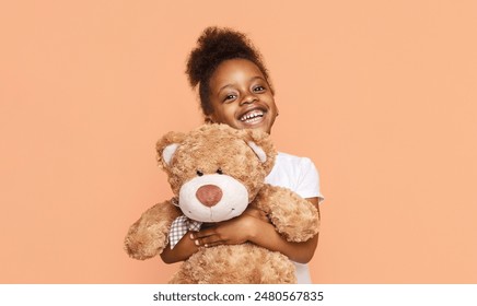 African American little girl with curly hair smiles while holding a brown teddy bear in front of a peach-colored background. She is wearing a white t-shirt and looks happy. - Powered by Shutterstock