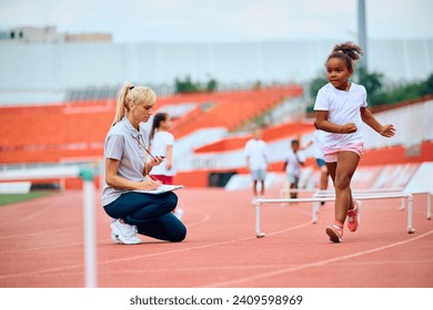 African American little girl running track with obstacles while coach is measuring time on stopwatch at the stadium. - Powered by Shutterstock