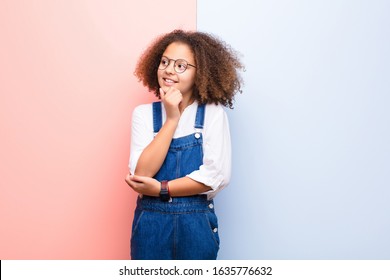 african american little girl smiling with a happy, confident expression with hand on chin, wondering and looking to the side against flat wall