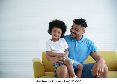 An African American Little Son Sat On His Father's Lap At Home. Dad Hug And Smile Looking At His Boy With Love On A White Background