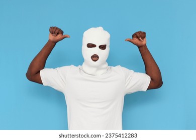 african american male bandit in white balaclava raises his hands and points to himself on blue isolated background, unrecognizable bully guy in mask shows biceps and gesture of strength