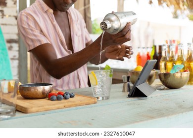 African american male bartender preparing cocktails using shaker at beach bar, unaltered. Small business, summer, refreshment and vacation. - Powered by Shutterstock