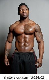 African American Male Body Builder Posing On A Gray Background While Looking Up And Wearing Black Shorts In A Studio Setting.