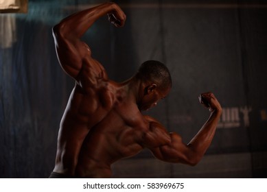 African American Male Body Builder Posing On A Black Background In Studio Setting. Back View