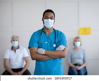 African American Male Doctor In Face Mask Posing With Crossed Arms And Looking At Camera In Clinic, Mature Patients Waiting For Covid-19 Vaccination On Background. Healthcare And Medicine Concept