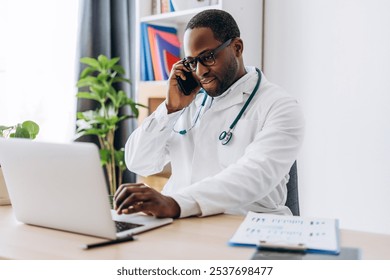 African American male doctor in glasses with stethoscope typing on laptop, talking on mobile phone. Healthcare, medicine concept - Powered by Shutterstock