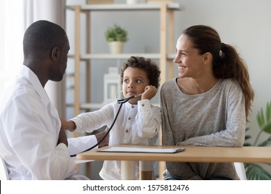 African american male doctor showing how phonendoscope works to small cute kid boy patient not to be afraid. Young mommy visiting trustful kind pediatrician with little son for medical check up.