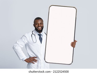 African American Male Doctor In White Coat With Stethoscope Holding Huge Phone With Blank Screen, Posing On Light Background. Medical App, Website, Therapist Advice, Health Care, Ad And Offer