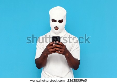 african american male hacker in white balaclava uses smartphone on blue isolated background, bully in mask is engaged in online crime on the phone