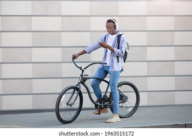 African American Male Hipster With Smartphone And Headset Riding Bike Near Brick Wall On Street, Copy Space. Positive Black Guy Listening To His Favorite Music During Bicycle Ride Outside