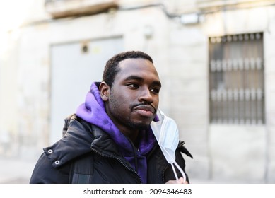 African American male in hoodie and outerwear taking off face mask and looking away on blurred background of building on city street