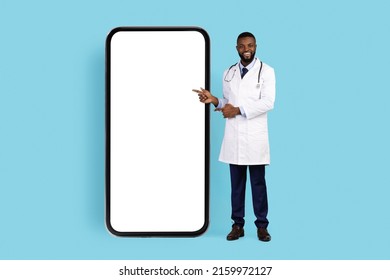 African American Male Physician In White Uniform Pointing Fingers At Big Phone Screen While Standing Over Blue Studio Background, Smiliing Black Doctor Standing Near Blank Cellphone, Mockup