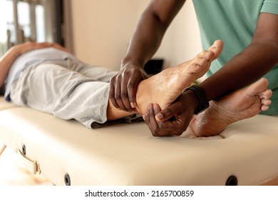 African american male physiotherapist giving foot massage therapy to caucasian senior woman at home. Physiotherapy and rehabilitation concept