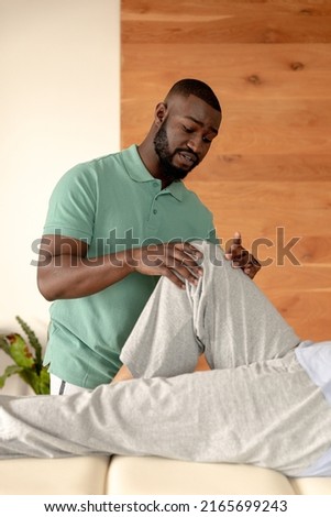 African american male physiotherapist giving leg massage therapy to caucasian senior woman at home. Physiotherapy and rehabilitation concept