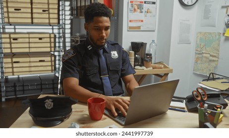 An african american male police officer analyzes evidence on a laptop in a crowded detective's office.