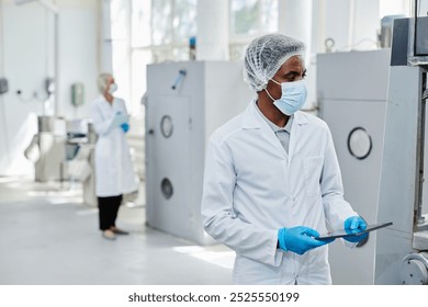 African American male process technician wearing protective mask and lab coat inspecting manufacturing machine while controlling production process at pharmaceutical factory, copy space - Powered by Shutterstock