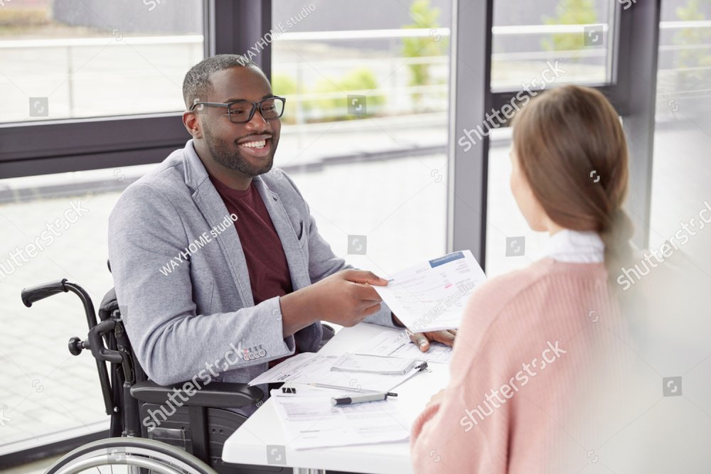 African American male sits at workplace in wheelchair, shares creative ideas and opinions with female worker, demonstrate new business project have informal meeting. Physically handicapped man