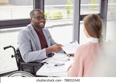 African American male sits at workplace in wheelchair, shares creative ideas and opinions with female worker, demonstrate new business project have informal meeting. Physically handicapped man