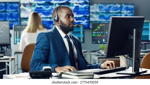 African American Male Stock Trader Or Broker Working At Stock Exchange Office Using Headset And Computer On Background Of Multiple Monitors Showing Data, Ticker Numbers And Graphs. 