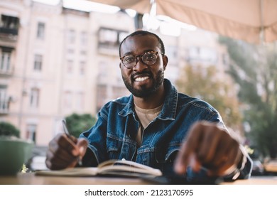 African American male student with education textbook for learning and studying in sidewalk cafe, cheerful hipster guy in classic glasses for eyes correction preparing for exams and smiling outdoors